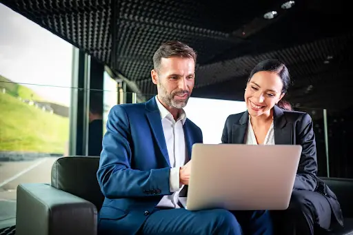 Two business professionals, a man and a woman in suits, sit on a couch smiling and looking at a laptop screen in a modern office with large windows.