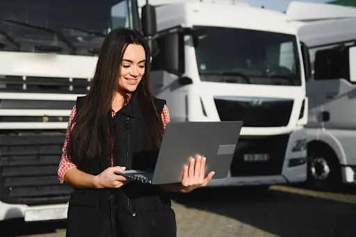 A woman stands outdoors in front of several white trucks, holding and looking at a laptop with a smile. She wears a checkered shirt and a black vest.