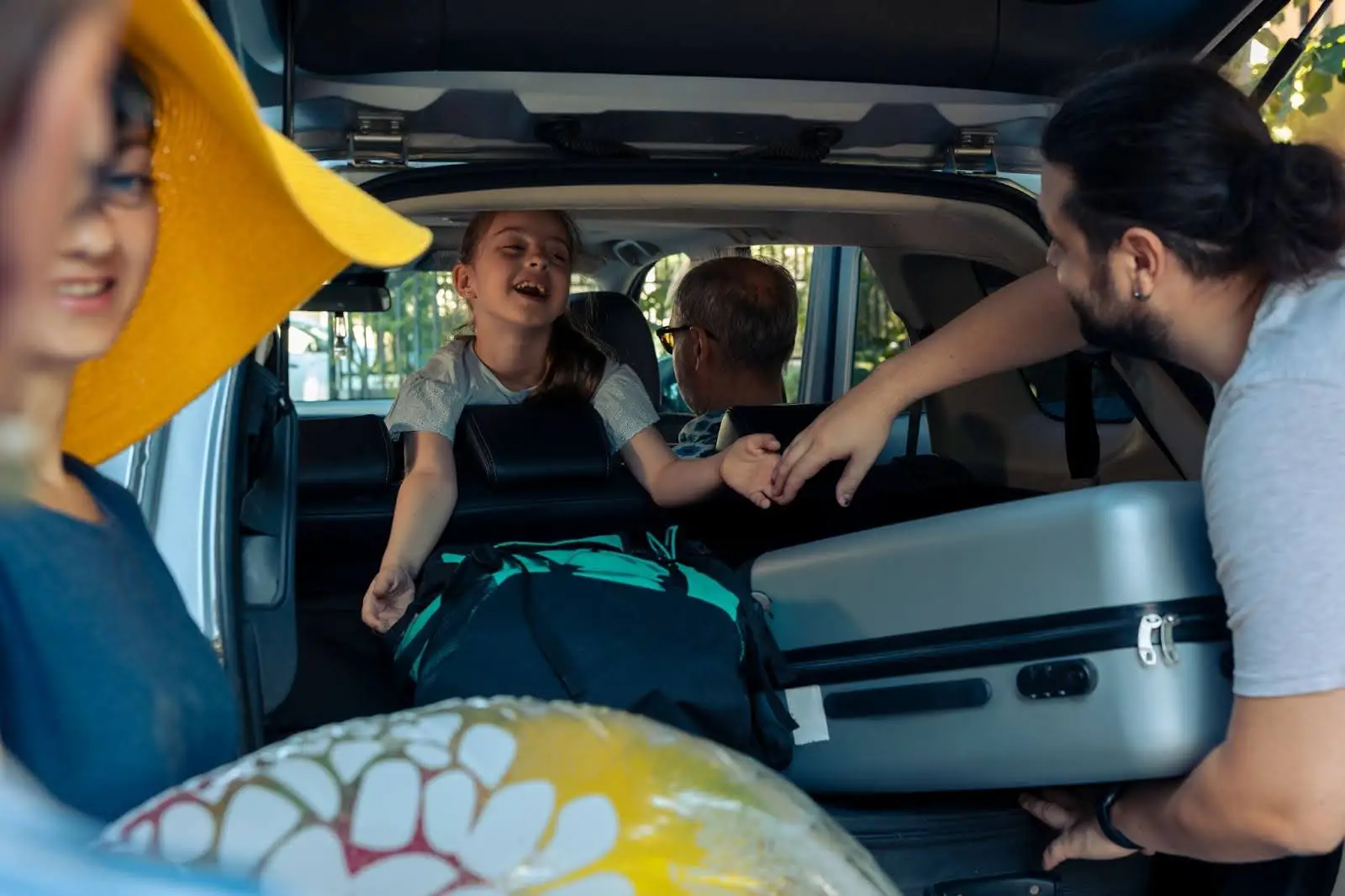 A family packs their car for a trip. A smiling girl sits in the back seat, reaching out, while adults load luggage and a beach ball into the trunk&mdash;just like a seamless journey managed with PerGO NEMT Software. Sunlight filters through the trees.