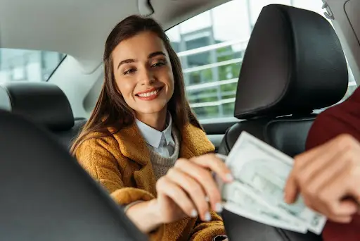 A woman sitting in a cars back seat smiles as she hands cash to the driver, who is partially visible in the front seat.