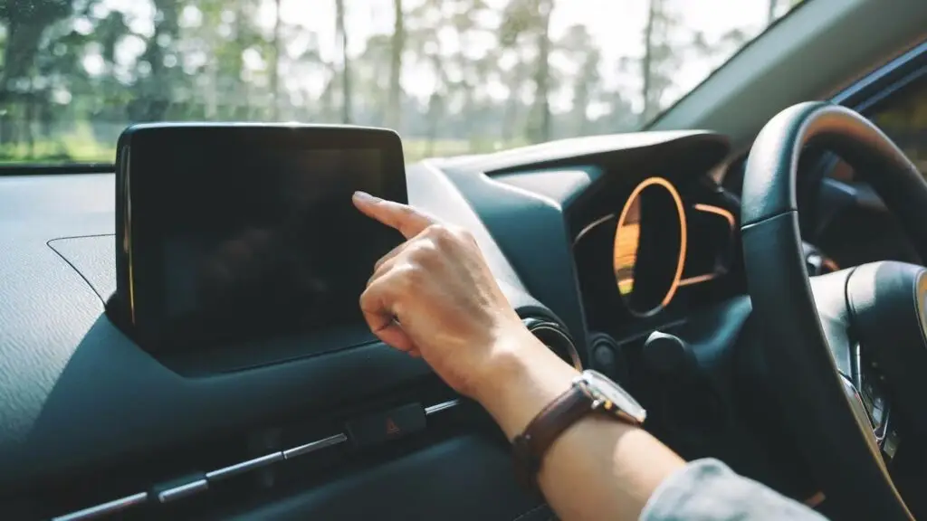 A person&rsquo;s hand is reaching out to touch the touchscreen display on a car dashboard, with sunlight shining through trees outside the vehicle.