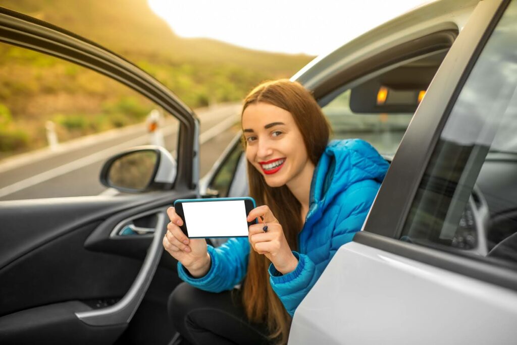 A smiling woman in a blue jacket sits in a car with the door open, holding a smartphone with a blank white screen toward the camera. The car is parked on a road with greenery in the background.