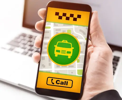 A hand holding a smartphone displaying a taxi app, with a map background, a yellow taxi icon, and a yellow Call button on the screen. A laptop keyboard is visible in the blurred background.