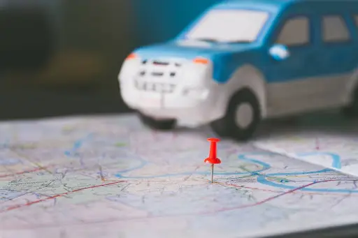 A small blue and white toy car sits on a paper map, marked by a red pushpin in the foreground. The focus is on the map and pin, evoking themes of travel and planning&mdash;ideal for showcasing PerGo transportation dispatch software.