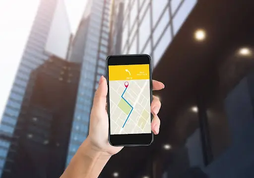 A hand holds a smartphone displaying a navigation map with a blue route line and a red location pin, showcasing PerGo transportation dispatch software against a backdrop of tall modern glass buildings.