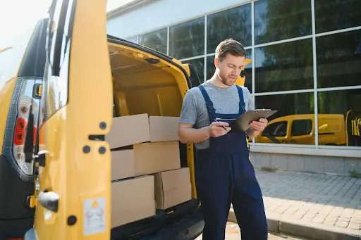 A delivery worker in blue overalls stands by an open yellow van filled with cardboard boxes, using PerGo transportation dispatch software on a clipboard outside a modern glass building.