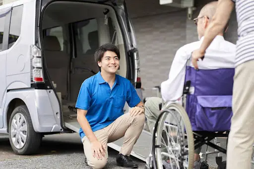 A caregiver in a blue shirt kneels and smiles at an elderly man in a wheelchair by a van with an open door and ramp, while another person stands nearby&mdash;made smoother with PerGo transportation management software.