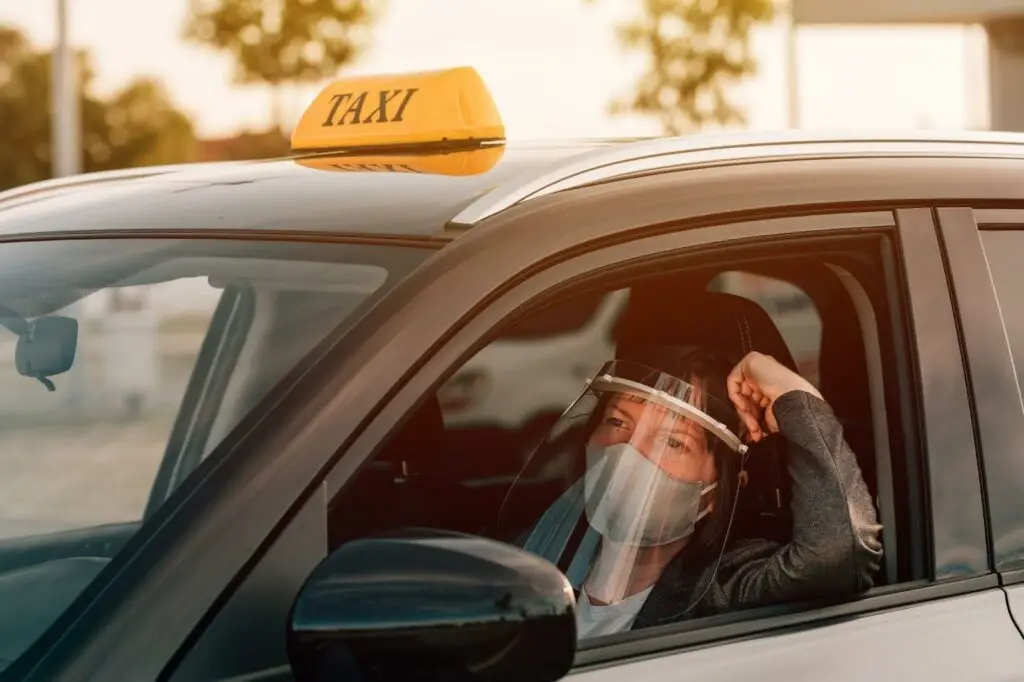 A person wearing a face mask and face shield sits in the driver's seat of a taxi, looking out the window with their arm resting on the door. The taxi sign is visible, highlighting how transportation dispatch software supports safe rides.