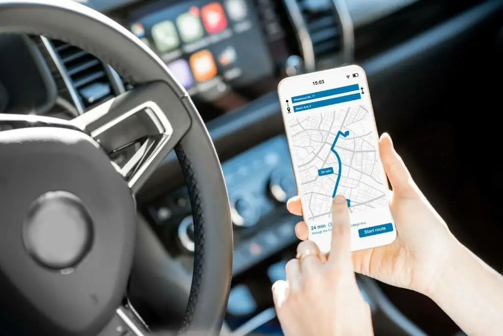 A person in a car uses a smartphone with a navigation app powered by PerGo transportation dispatch software. The car’s steering wheel and dashboard are visible in the background.
