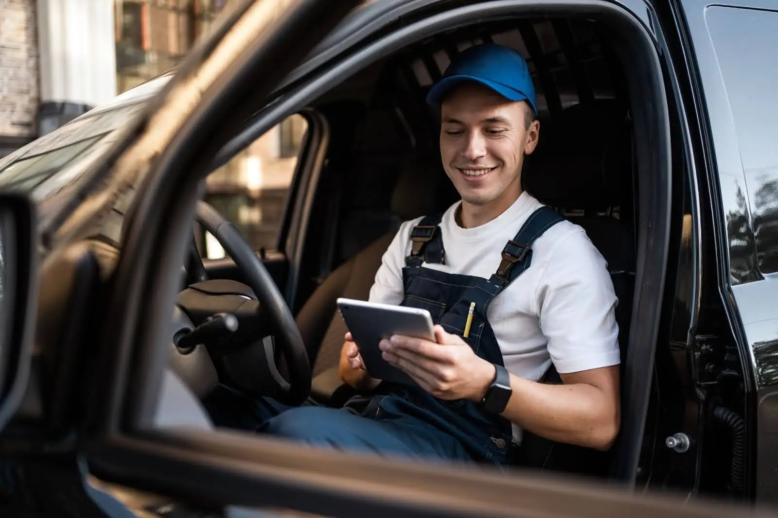 A smiling man in a blue cap and overalls sits in the driver&rsquo;s seat of a vehicle, looking at a tablet device in his hands, using transportation dispatch software. The driver&rsquo;s side door is open as he checks important information for his route.