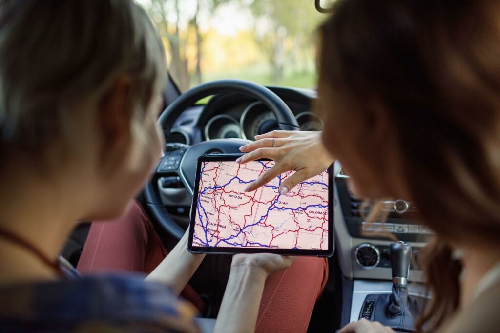 Two people sitting in a car look at a tablet displaying a map with highways and routes. Using PerGo transportation dispatch software, they plan their trip as one holds the tablet while the other points to a location on the screen.