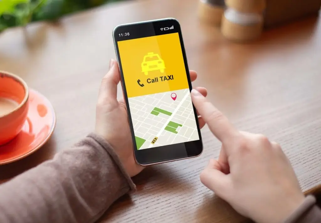 A person holds a smartphone displaying the PerGo taxi booking app with a map and a Call TAXI button, sitting at a wooden table&mdash;highlighting modern transportation dispatch software in action.