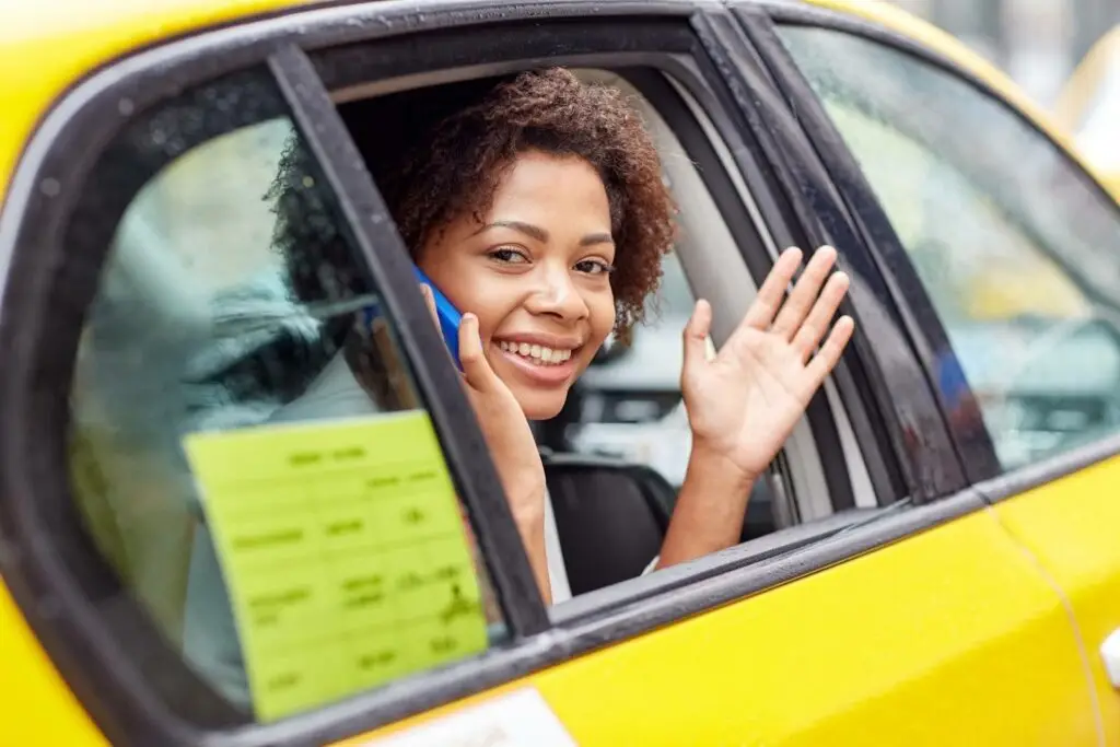 A smiling person sits in the back seat of a yellow taxi, holding a phone to their ear and waving out the open window&mdash;showcasing how transportation dispatch software streamlines every ride. A yellow document is visible on the car door.