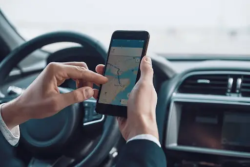 A person sitting in a car uses the PerGo transportation dispatch software on a smartphone, holding the phone with one hand and touching the screen with the other. The steering wheel and dashboard are visible.