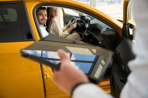A man and woman sit in the front seats of a yellow car, smiling at a person holding a tablet outside the vehicle&mdash;perhaps demonstrating innovative NEMT software in a modern car dealership or showroom.