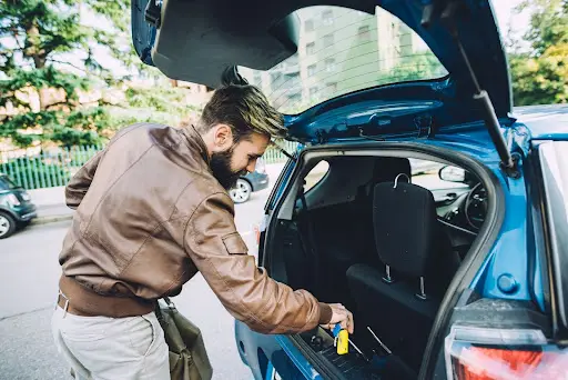 A man with a beard and a brown leather jacket places items into the open trunk of a blue car parked on a street, illustrating efficient organization much like transportation management software. Trees and buildings are visible in the background.