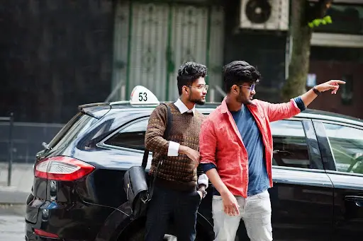 Two men stand beside a black car with a taxi sign on its roof, discussing directions. Their engaged interaction hints at efficient coordination&mdash;perhaps aided by transportation dispatch software&mdash;on the busy city street.