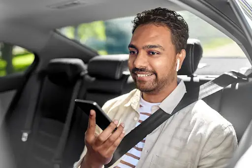 A man sits in the backseat of a car, wearing a seatbelt and wireless earbuds, smiling as he speaks into his smartphone. Sunlight streams through the window&mdash;seamless travel like this is possible with advanced transportation dispatch software.