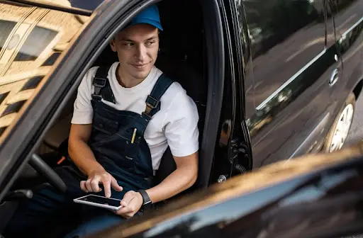 A person wearing a white shirt, blue overalls, and a blue cap sits in the driver's seat of a car, smiling and using a tablet with PerGo transportation dispatch software, as a city street is reflected in the car’s window.