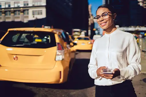 A woman wearing glasses and a white blouse stands smiling on a city street, holding a smartphone, with yellow taxis and tall buildings in the background—perfectly capturing the spirit of PerGo transportation dispatch software.