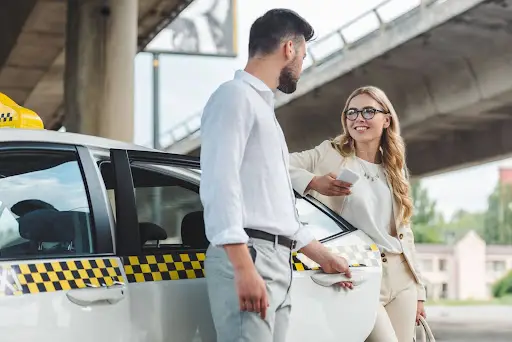A man holds open a taxi door for a smiling woman standing nearby. Under an overpass, they chat as the woman, in business attire, checks her smartphone—possibly coordinating her ride with transportation dispatch software.