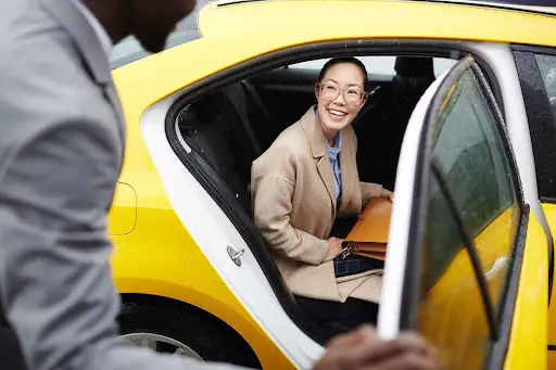 A smiling woman with glasses sits in the backseat of a yellow taxi, holding an orange folder, as a man opens the car door for her&mdash;seamlessly coordinated with PerGo transportation dispatch software.