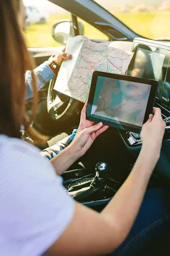 Two people sit in a car; one holds a paper map while the other uses a tablet displaying PerGo transportation management software, both navigating their route together.
