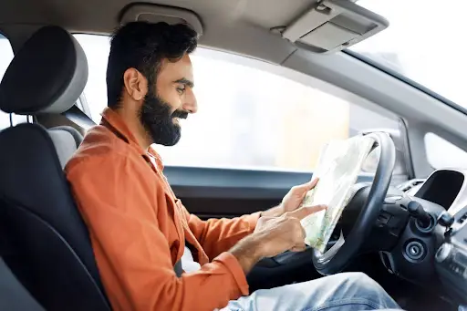 A man sits in the driver&rsquo;s seat of a car, smiling as he looks at and points to a paper map, appearing to navigate or plan a route&mdash;just like effective transportation dispatch software such as PerGo streamlines trip planning.