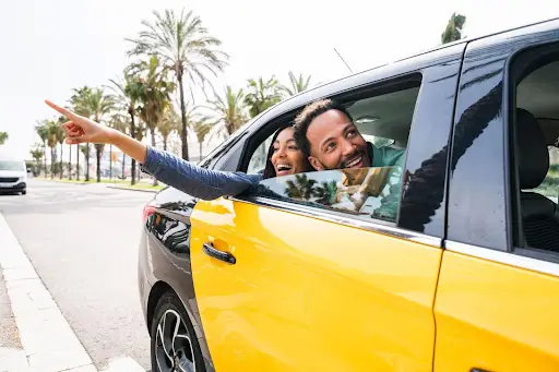 A smiling man and woman lean out of the window of a yellow and black taxi, enjoying the moment as the woman excitedly points at something outside. Palm trees line the sunny street&mdash;PerGo transportation dispatch software keeps their ride seamless.