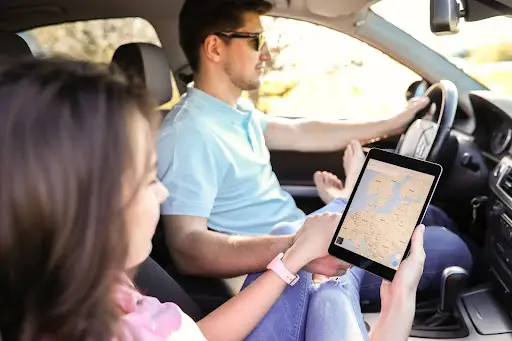A woman in the passenger seat of a car holds a tablet displaying PerGo transportation dispatch software. A man drives the car, focusing on the road ahead as sunlight streams through the windows.