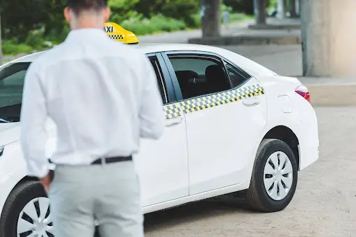 A person in a white shirt stands near a white taxi with a yellow taxi sign on its roof, parked on a street&mdash;offering a glimpse into efficient transportation dispatch software like PerGo. The person's back is facing the camera.