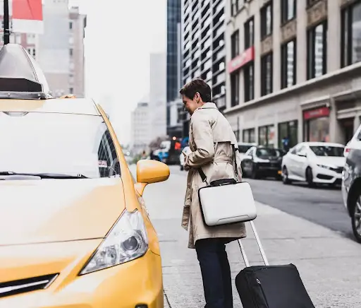 A person with short hair in a beige trench coat stands on a city street next to a yellow taxi, holding a rolling suitcase and shoulder bag&mdash;ready to get into the cab, perhaps aided by PerGo&rsquo;s advanced transportation dispatch software.