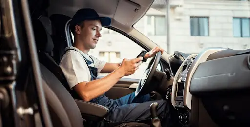 A man wearing a cap and work overalls sits in the driver’s seat of a van, holding the steering wheel with one hand and checking transportation dispatch software on his smartphone with the other.