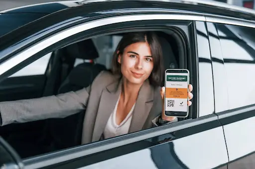 A woman sitting in a car holds out her smartphone, displaying a digital vaccine passport with a QR code. She smiles slightly, ready to show her proof of vaccination—making travel and Transportation management Software like PerGo more efficient than ever.