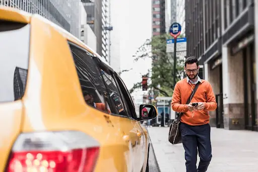 A man wearing glasses, an orange sweater, and carrying a bag walks on a city sidewalk, looking at his phone&mdash;perhaps checking PerGo transportation dispatch software&mdash;as a yellow taxi waits at the curb amid tall office buildings.