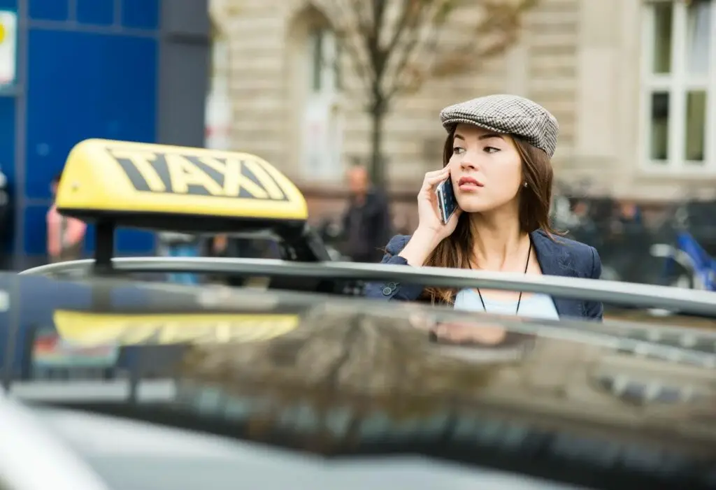 A young woman in a flat cap stands by a taxi, talking on her phone, with a city street and blurred buildings in the background—perhaps coordinating her ride with transportation dispatch software.