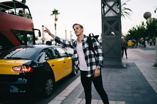 A man in a black and white plaid shirt with a backpack stands on a city sidewalk, raising his arm to hail a yellow and black taxi&mdash;an everyday scene made more efficient with transportation dispatch software. A red double-decker bus and palm trees are in the background.