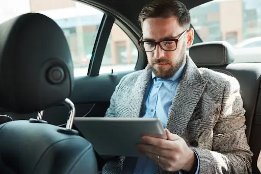 A man with glasses and a beard, wearing a grey plaid blazer and blue shirt, sits in the backseat of a car concentrating on his tablet, reviewing details on PerGo transportation dispatch software.