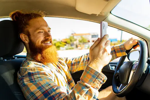 A smiling man with a red beard and plaid shirt is driving a car, holding the steering wheel with one hand and taking a selfie with his phone in the other&mdash;perhaps showing off his ride using PerGo NEMT Software. Sunlight shines through the window.
