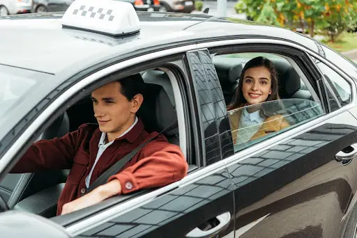 A woman sits in the back seat of a black taxi, smiling, while the male driver focuses on the road—efficiently navigating with PerGo transportation dispatch software. Trees and parked cars pass by outside.