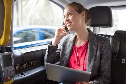 A woman in business attire sits in the back seat of a car, smiling while talking on her phone and holding a laptop&mdash;efficiently managing tasks with PerGo transportation dispatch software.