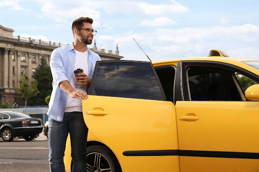 A smiling man holding a coffee cup stands beside an open door of a yellow taxi on a city street, showcasing how PerGo transportation dispatch software streamlines urban travel, with buildings and cars in the background.