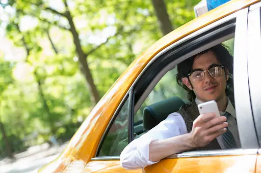 A person wearing glasses sits in the driver's seat of a yellow taxi, holding a smartphone and looking out the open window&mdash;perhaps using transportation dispatch software. The background is blurred with green trees and sunlight.