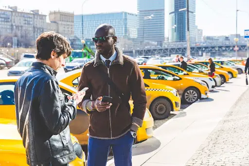 Two men stand on a city sidewalk near a row of parked yellow taxis. One checks his phone, while the other in sunglasses uses his device&mdash;perhaps managing rides with PerGo&rsquo;s advanced Transportation Management Software. Tall buildings rise in the background.