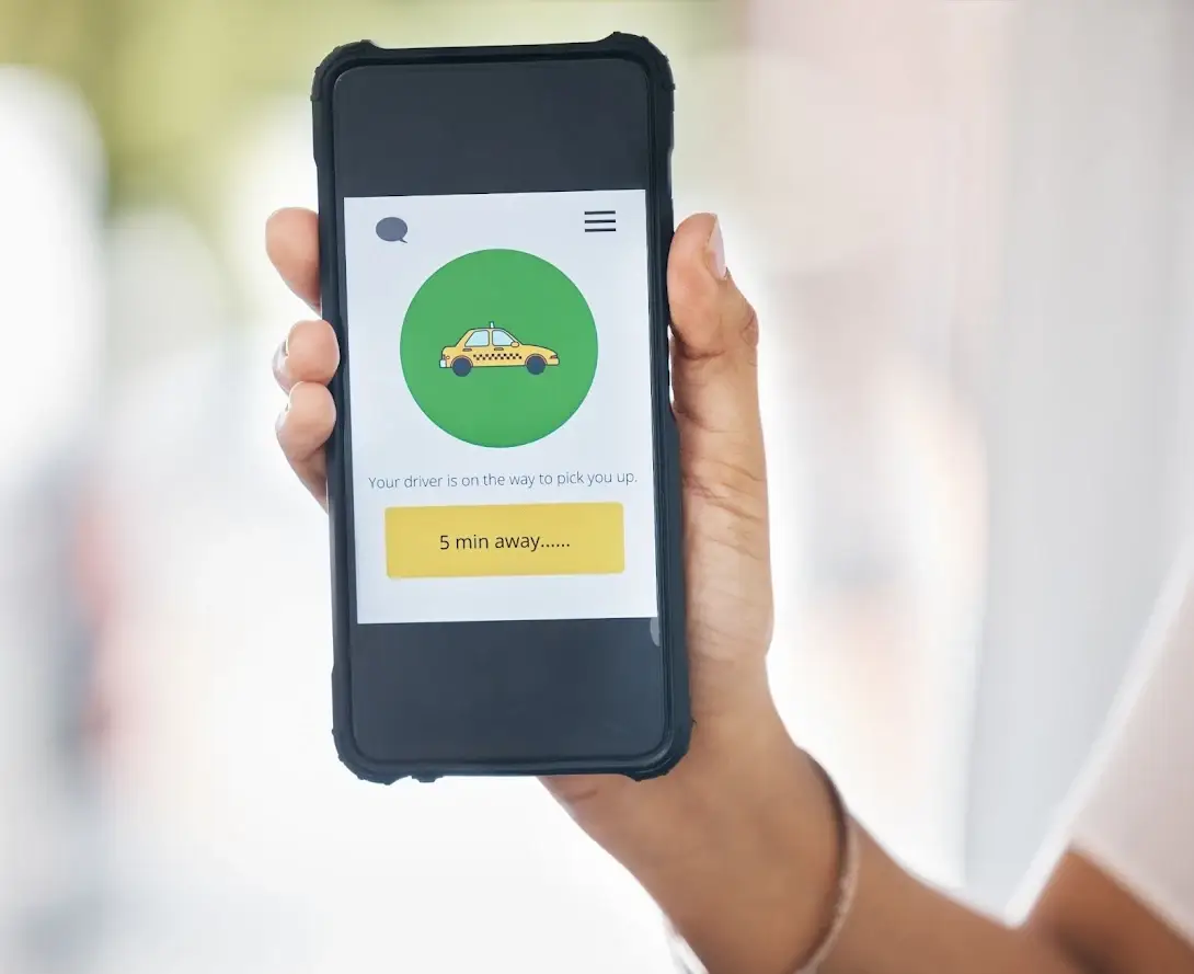 A hand holding a smartphone displaying a taxi app screen with a graphic of a yellow cab and text stating, &ldquo;Your driver is on the way to pick you up. 5 min away.&rdquo;.