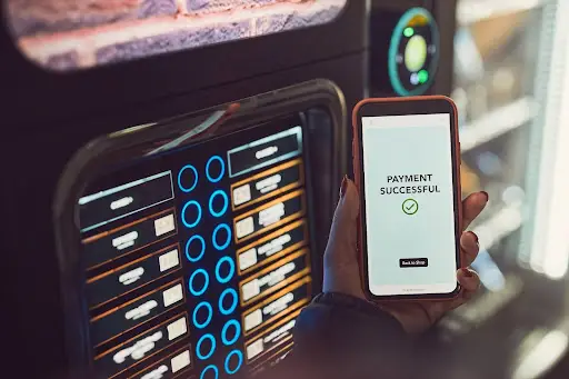 A person holds a smartphone showing a “Payment Successful” message with a checkmark in front of a vending machine, indicating a contactless or online payment.