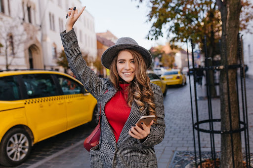 A young woman in a gray hat and plaid coat smiles while hailing a yellow taxi, using PerGo transportation management software on her smartphone. She stands near trees and parked cars on a sunny day in the city.